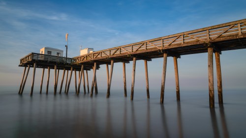 A wooden pier extends over calm water, with a small building at the end and a clear blue sky above.