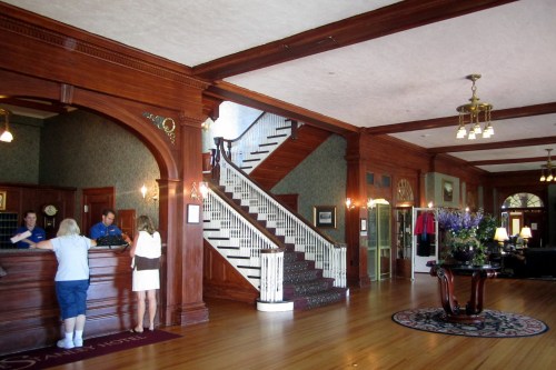 A spacious hotel lobby with wooden decor, a staircase, and guests checking in at the reception desk.