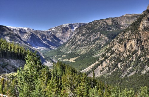 A scenic mountain valley with lush green trees, rocky cliffs, and snow-capped peaks under a clear blue sky.