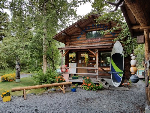 A rustic log cabin storefront surrounded by greenery, flowers, and a surfboard, with a wooden bench in front.
