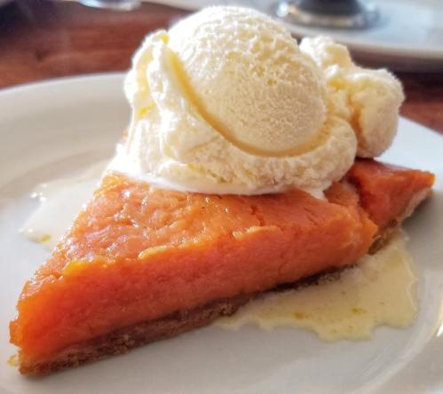 A slice of sweet potato pie topped with a scoop of vanilla ice cream on a white plate.