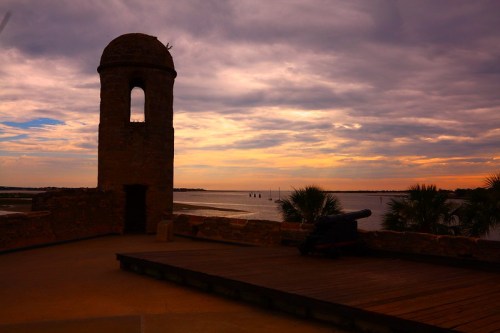Silhouette of a stone tower and cannon against a colorful sunset over a calm waterway.