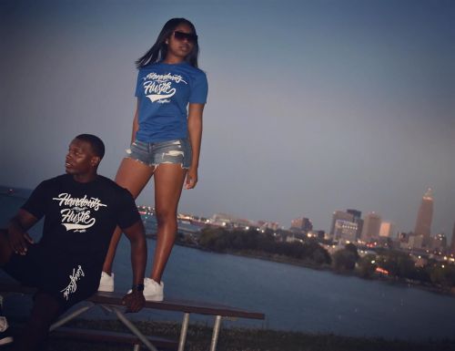 A man in a black shirt sits on a bench, while a woman in a blue shirt stands beside him, with a city skyline in the background.