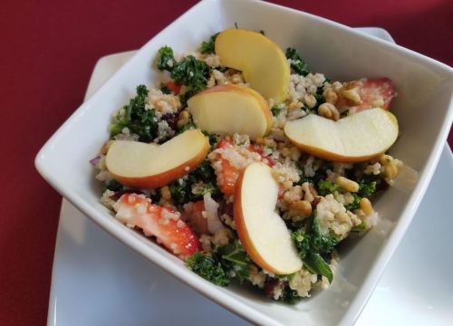 A bowl of quinoa salad with kale, strawberries, walnuts, and apple slices on top, served on a white plate.