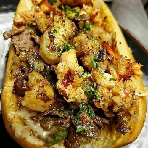 A loaded sandwich topped with grilled shrimp, beef, and fresh herbs on a toasted bun.