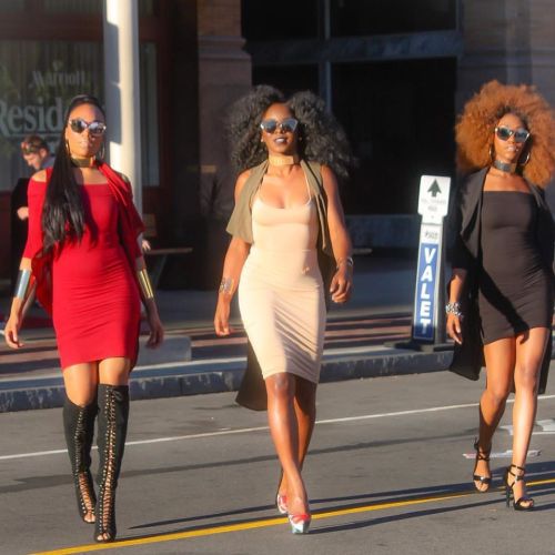 Three women confidently walk down the street, showcasing stylish outfits and bold accessories in a vibrant urban setting.