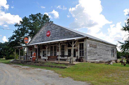 A rustic, weathered general store with a Gulf sign, surrounded by greenery and a gravel path under a blue sky.