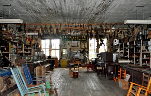 A vintage store interior filled with old tools, furniture, and memorabilia, featuring shelves and rustic decor.