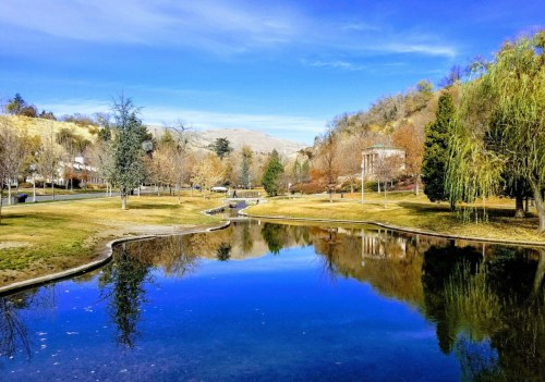 A serene park scene with a calm pond reflecting trees and blue sky, surrounded by grassy areas and distant hills.