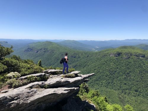 A person stands on a rocky ledge, overlooking a lush green mountain landscape under a clear blue sky.