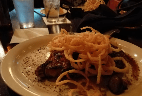 A plate of grilled meat topped with crispy onion rings, served on a table with drinks and side dishes in the background.