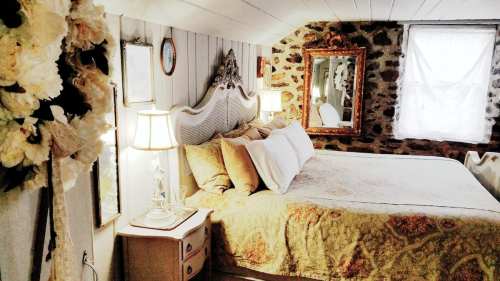 Cozy bedroom with a large bed, decorative pillows, vintage mirrors, and stone walls, illuminated by soft lamps.