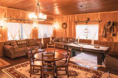 Cozy wooden interior with a pool table, seating area, and rustic decor, featuring warm lighting and large windows.