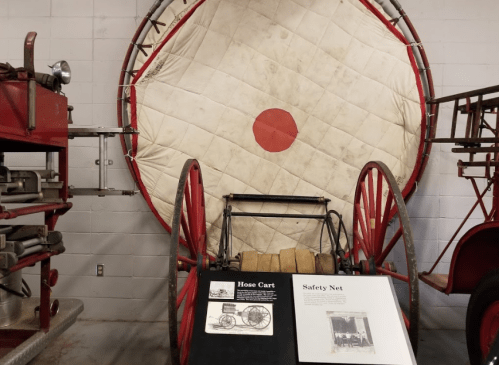 A vintage hose cart displayed in front of a large safety net with a red circle, in a museum setting.