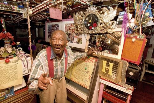 An elderly man gestures animatedly in a colorful, eclectic room filled with decorations and memorabilia.
