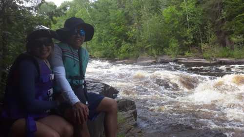 A couple sits on a rocky riverbank, wearing life jackets and hats, with a rushing river and lush greenery in the background.