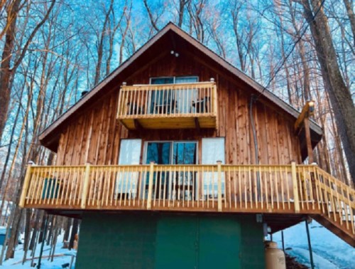 A wooden cabin with balconies, surrounded by trees and snow, set against a clear blue sky.
