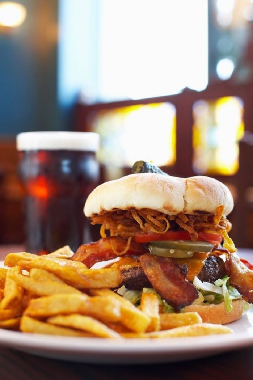 A towering burger topped with crispy onions, bacon, and pickles, served with golden fries and a glass of dark soda.