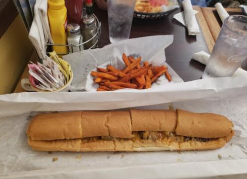 A long sandwich filled with toppings, served with a side of sweet potato fries and condiments on a table.