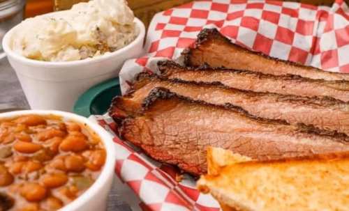 A plate featuring sliced brisket, baked beans, and a side of potato salad, all served with checkered paper.