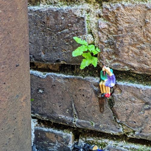 A small figurine of a couple sitting on a brick wall, with a green plant growing nearby.