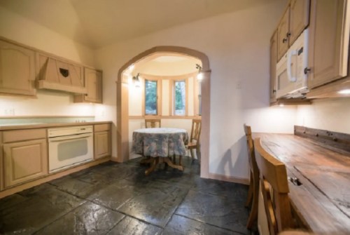 Cozy kitchen with a round table, wooden chairs, and stone floor, featuring an archway and natural light from windows.