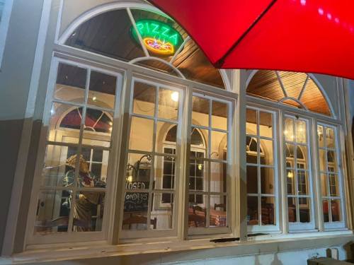 A view of a pizza restaurant through large windows, with a neon sign and a red umbrella outside.