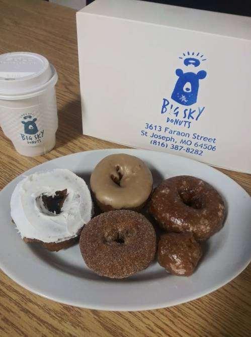 A plate of assorted donuts next to a coffee cup and a Big Sky Donuts box on a wooden table.