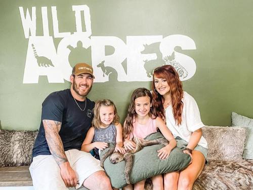 A family poses with a sloth on a couch in front of a green wall with "Wild Acres" painted on it.