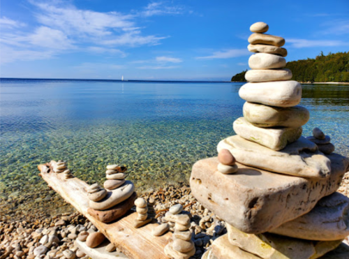 A serene lakeside scene with stacked stones on a log, clear water, and a blue sky in the background.