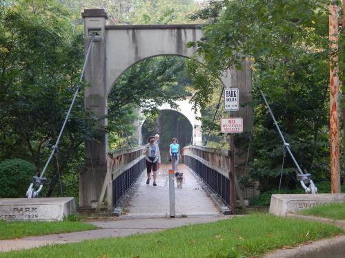Two people walk a dog across a bridge in a park, surrounded by lush greenery and signs indicating park rules.