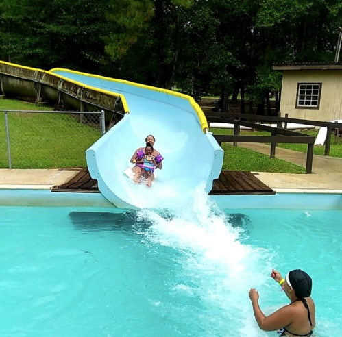 A person slides down a water slide into a pool while another person splashes water nearby.