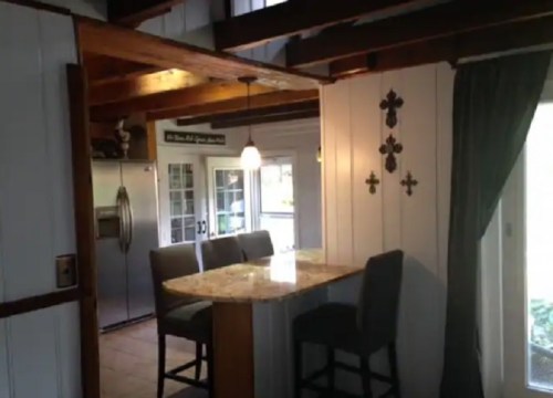 Cozy kitchen area with a granite countertop, bar stools, and wooden beams, featuring a window and stainless steel fridge.