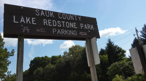Sign for Sauk County Lake Redstone Park, indicating parking to the left, with trees and blue sky in the background.