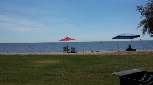 A beach scene with a red umbrella, green chair, and a blue tent by the water under a clear sky.