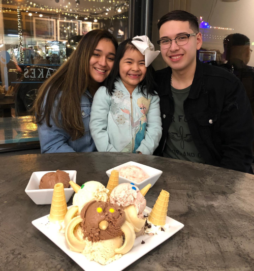 A girl with a bow smiles between two young adults, with colorful ice cream desserts in front of them on a table.
