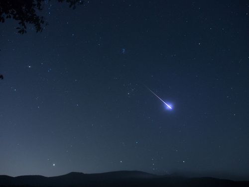 A night sky filled with stars, featuring a bright meteor streaking across the scene above dark silhouettes of mountains.