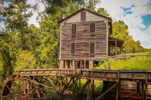 A rustic wooden mill with a waterwheel, surrounded by lush greenery and blue skies.
