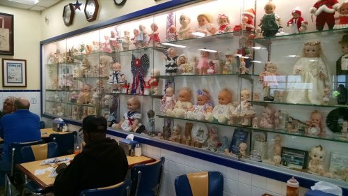 A wall display of vintage dolls in a diner, with patrons seated at tables below.