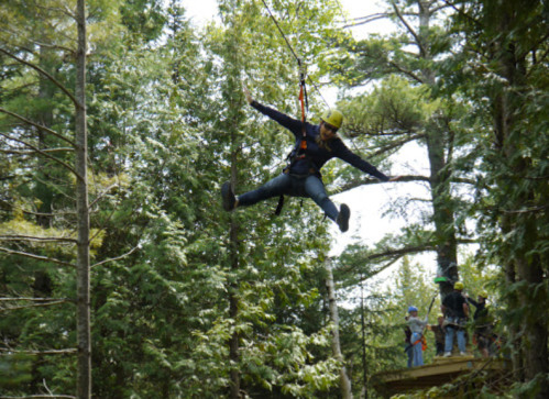 A person zip-lining through trees, arms outstretched, with others watching from a platform nearby.