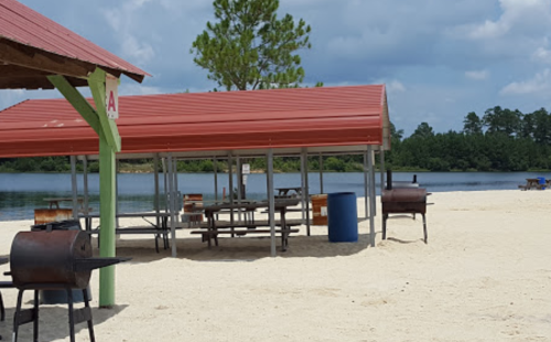 Beach scene with red-roofed pavilions, picnic tables, and grills near a calm lake surrounded by trees.