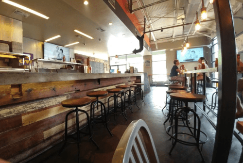 Interior of a casual restaurant with wooden bar stools, a counter, and patrons dining in a bright, open space.