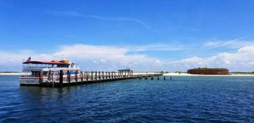A boat docked at a pier with a historic fort in the background, under a clear blue sky and calm waters.