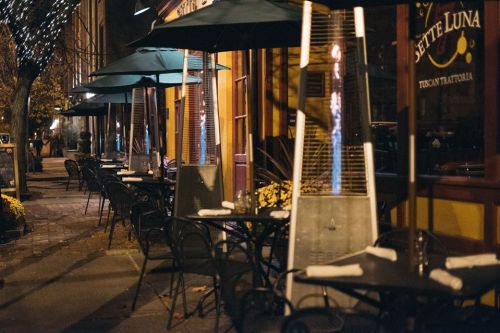 Outdoor dining area with empty tables, green umbrellas, and a heater, illuminated by soft evening lights.