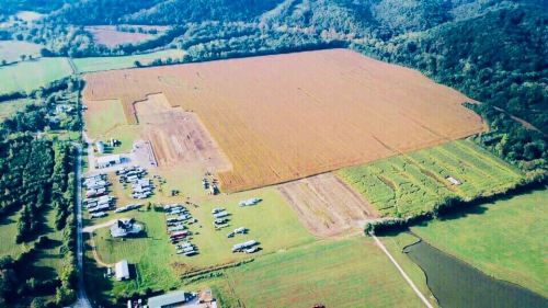 Aerial view of a large, golden field surrounded by green hills, with parked vehicles and a small building in the foreground.