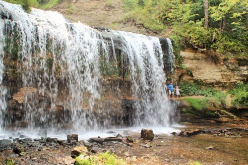 A scenic waterfall cascades over rocks, with two people standing nearby, surrounded by lush greenery.