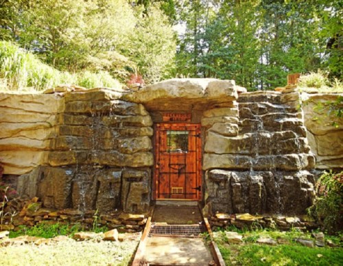 A wooden door set into a rocky, natural stone wall with water cascading down, surrounded by greenery.