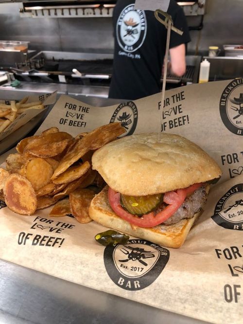 A delicious burger with pickles and tomatoes on a bun, served with crispy potato chips, all on branded paper.