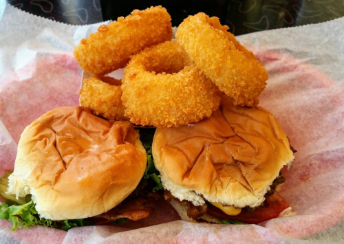 Two burgers with lettuce and tomato, topped with a stack of golden, crispy onion rings on a pink paper-lined tray.