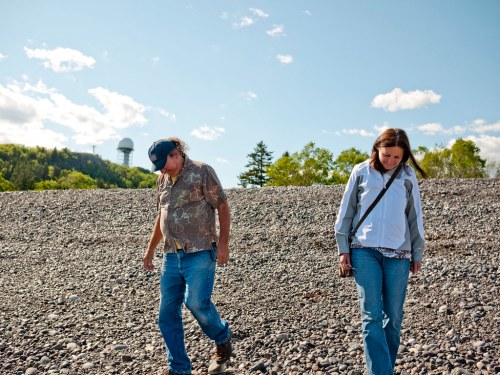 Two people walking on a rocky surface under a clear blue sky, with trees and a radar tower in the background.